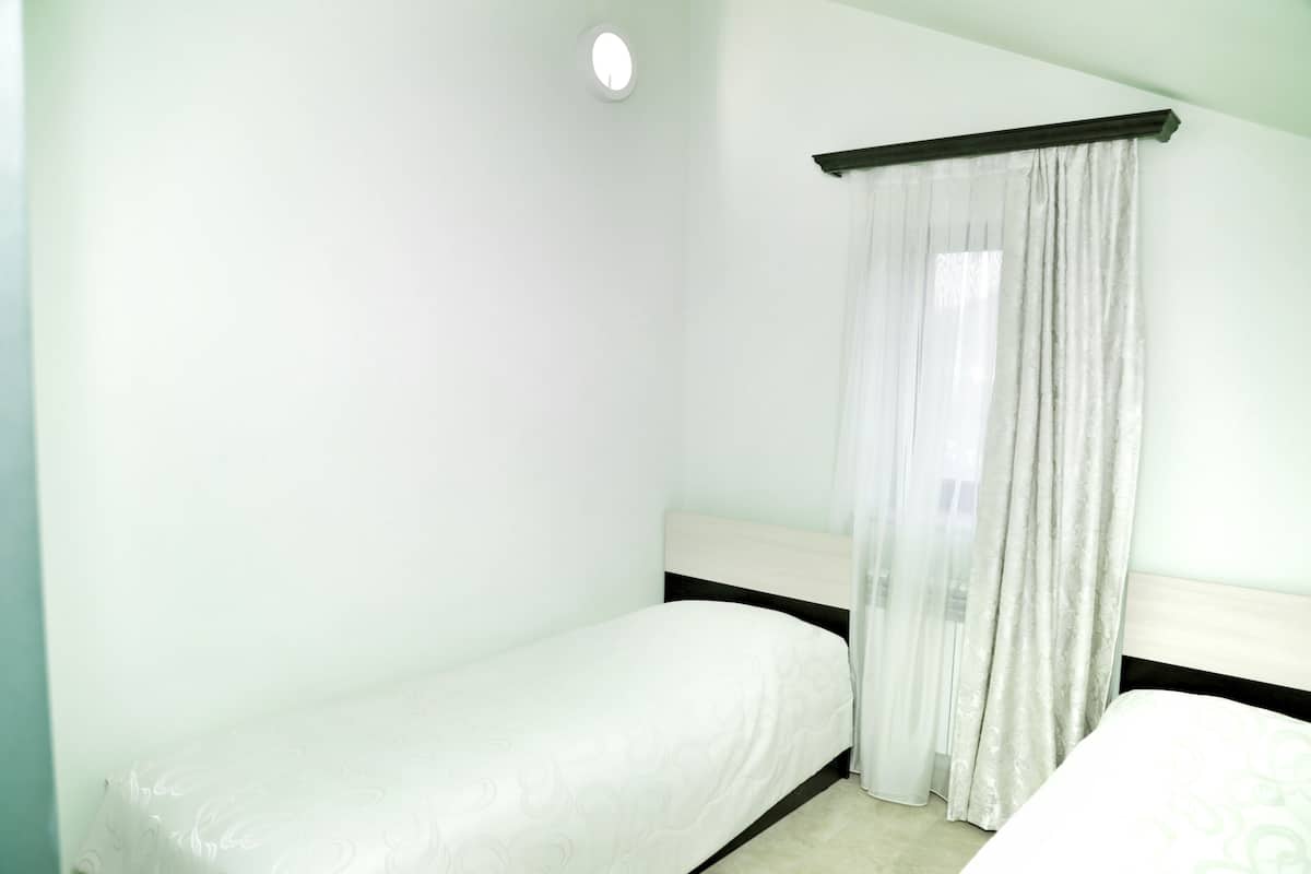 A small, clean room with two single beds, a window with sheer curtains, and a white wall.