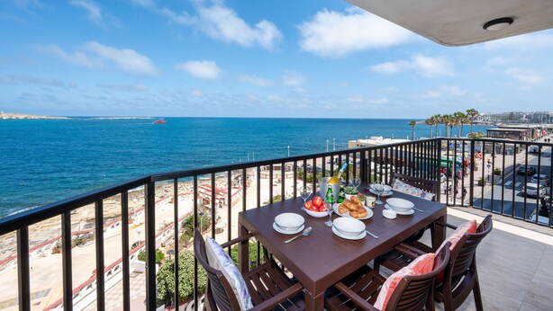 A balcony with a table set for two, overlooking a beach and the sea. - Islet Seafront Apartment Corner Terrace (St. Paul's Bay)