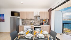 A modern kitchen with a dining area, equipped with a refrigerator, stove, and sink, and a large window offering a scenic view. - Islet Seafront Apartment Corner Terrace (St. Paul's Bay)