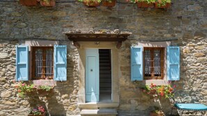 Ein Steinhaus mit einer zentralen Tür, flankiert von Fenstern mit blauen Fensterläden, und üppigen Pflanzgefäßen mit roten Blüten.