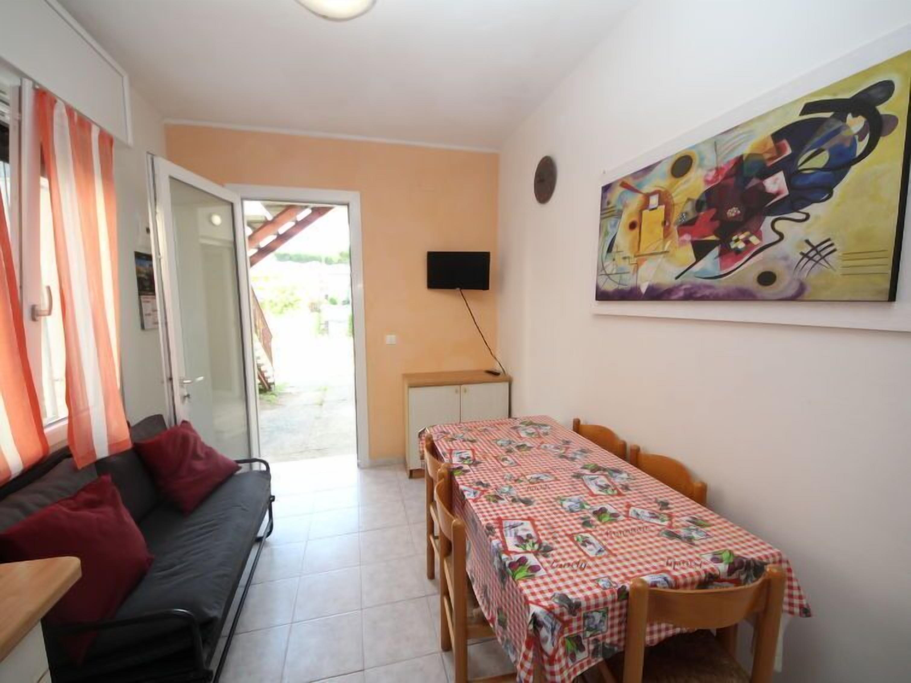 Un espace de vie compact comprenant un canapé, une table à manger recouverte d’une nappe à carreaux, un téléviseur fixé au mur et un tableau abstrait aux couleurs vives.