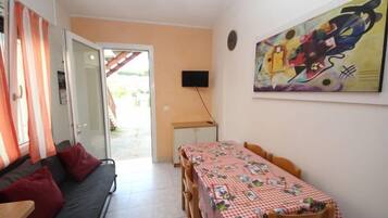 Ein kompakter Wohnbereich mit einer Couch, einem Esstisch mit kariertem Tischläufer, einem an der Wand befestigten Fernseher und einem farbenfrohen, abstrakten Gemälde.