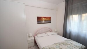 A bedroom with a bed, a nightstand, a drawer, a painting of a sunset, and a window with curtains.