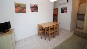 A small, well-lit room with a wooden dining table and chairs, a television on a cabinet, and artwork on the walls.