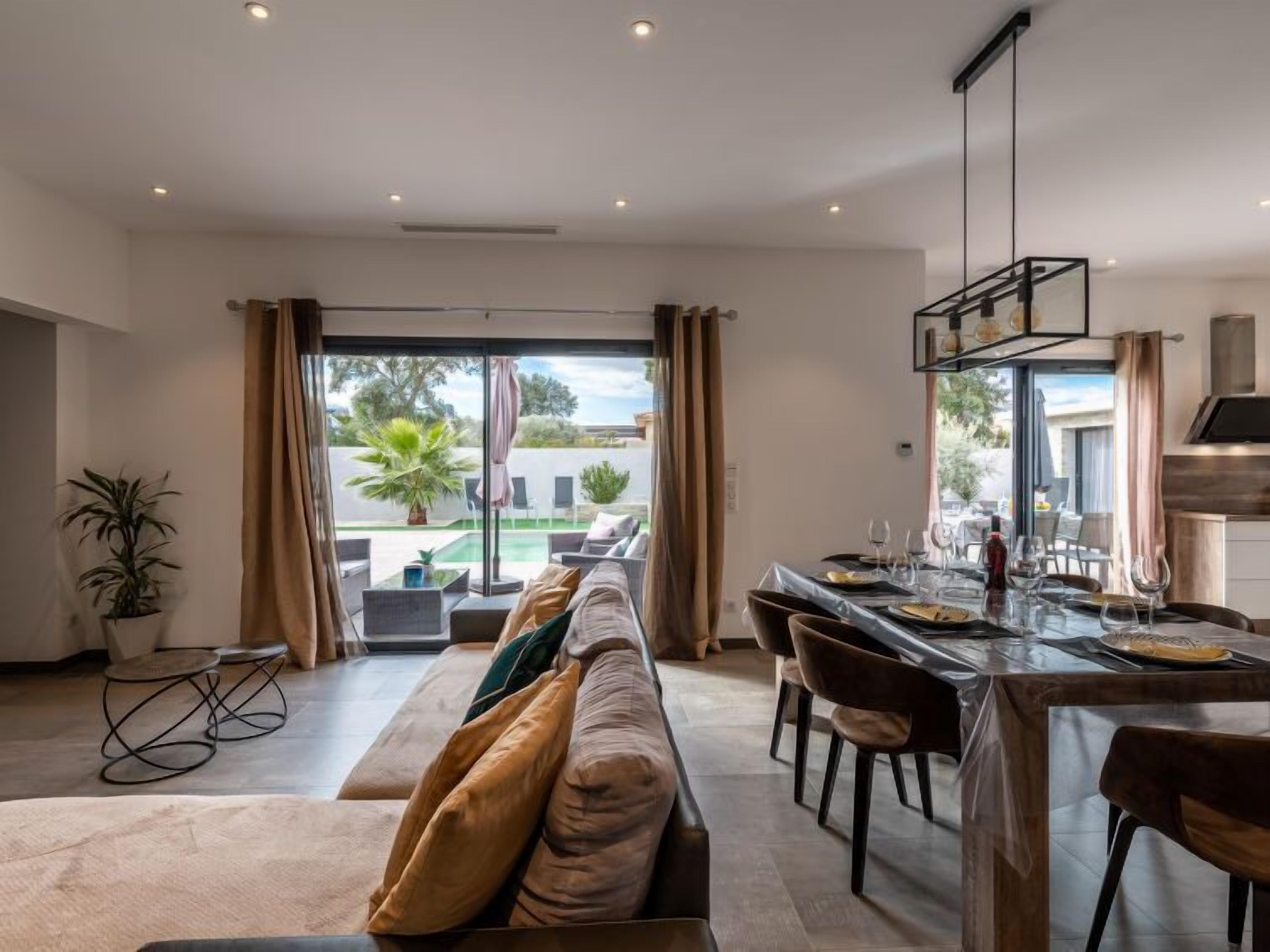 A modern kitchen with a dining area, large windows, and a view of a pool and greenery.