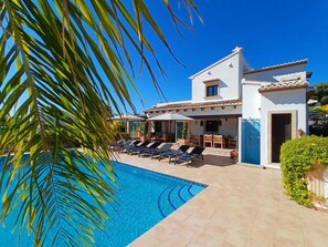 Un espace aménagé au bord de la piscine, avec des chaises longues, une terrasse et un bâtiment blanc doté d’un toit en tuiles.