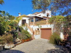 Une maison à deux étages avec un escalier en pierre menant à un balcon, entourée d’une végétation luxuriante et surplombant un ciel bleu azur.