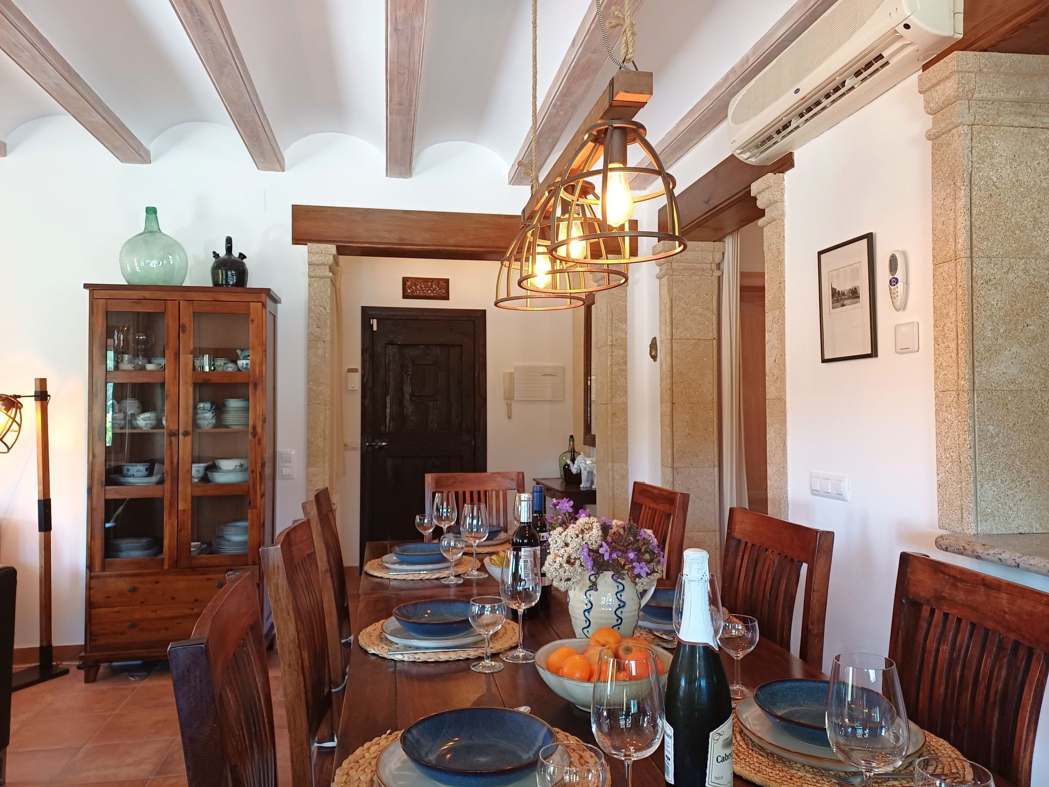 A dining area with a wooden table set for six, surrounded by chairs, with a cabinet of dishes and a hanging light fixture above.