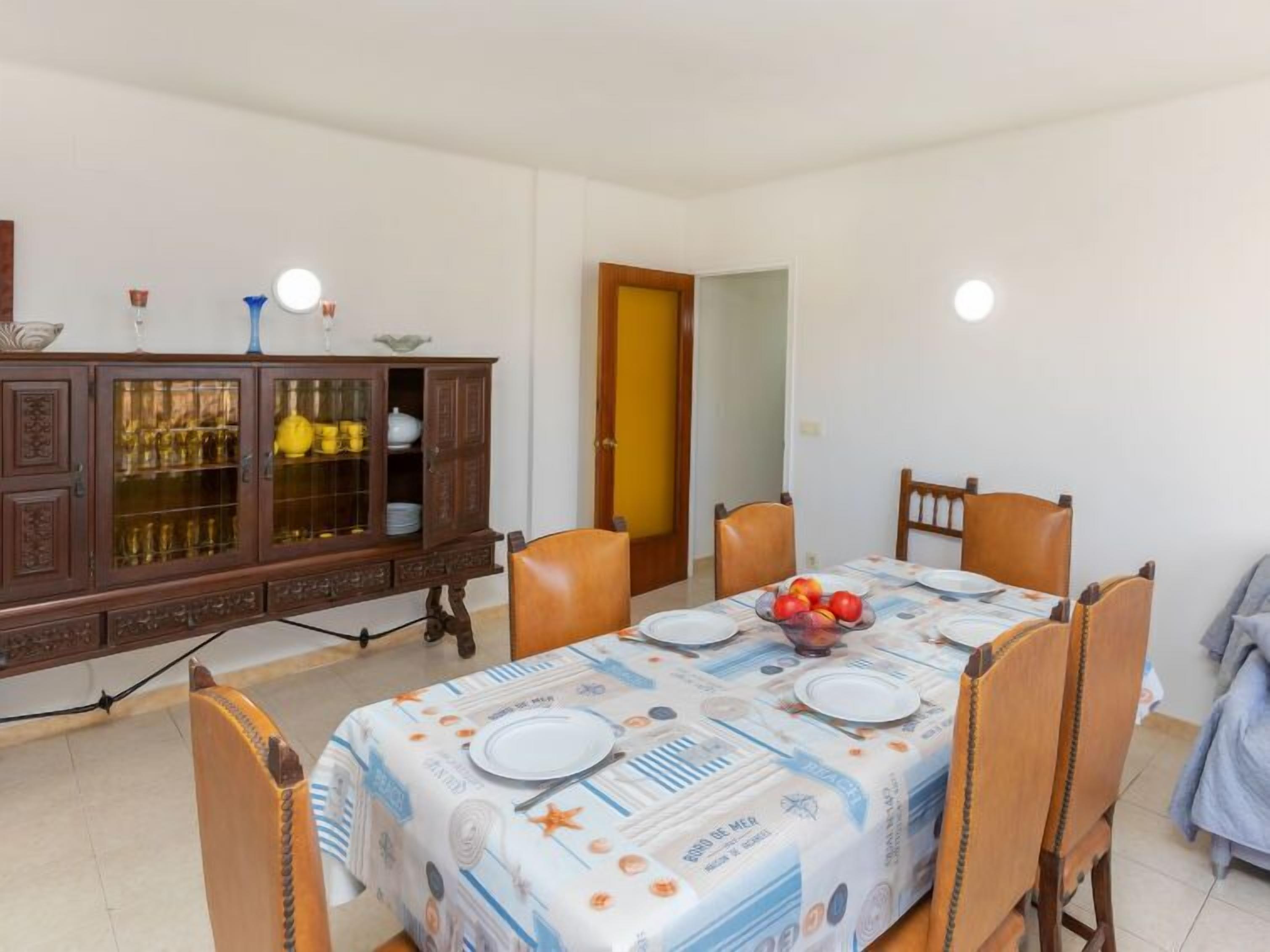 A dining area with a table set for four, surrounded by wooden chairs. A wooden cabinet with glass doors is to the left, containing various items.