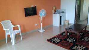 A room with a white plastic chair, a standing fan, a TV mounted on the wall, a small table, and a patterned rug. - Sandai resort (Marigat)