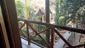 Un balcon doté d’une balustrade en bois, donnant sur un jardin où poussent différentes espèces de plantes, et sur une construction dont le toit est fait de chaume.