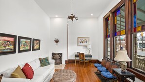 A living room with a white sofa, ottoman, and blue chairs, a fireplace, a dining table with chairs, and large windows with colorful stained glass. - Luxurious King Room in Historic Boutique Inn (Winston-Salem)