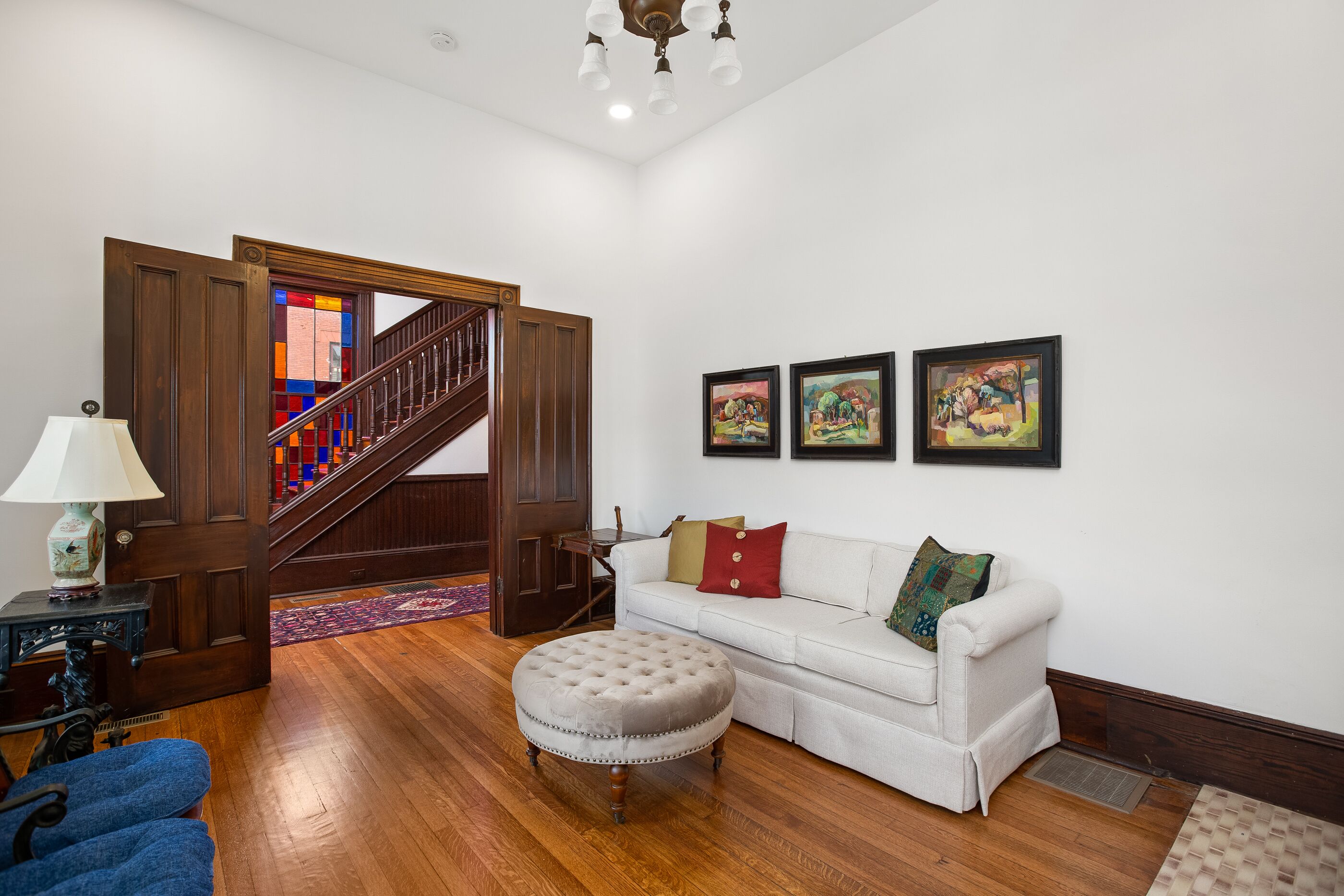 A living room with a wooden staircase, a white sofa, an ottoman, and three framed artworks on the wall.