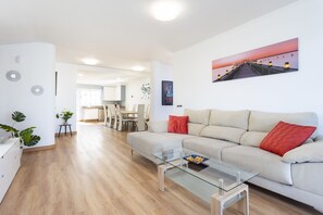A modern living room with a glass coffee table, a beige sofa with red cushions, and a dining area in the background.