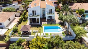 Une maison moderne à deux étages, dotée d’une piscine, entourée d’une végétation luxuriante et de palmiers.