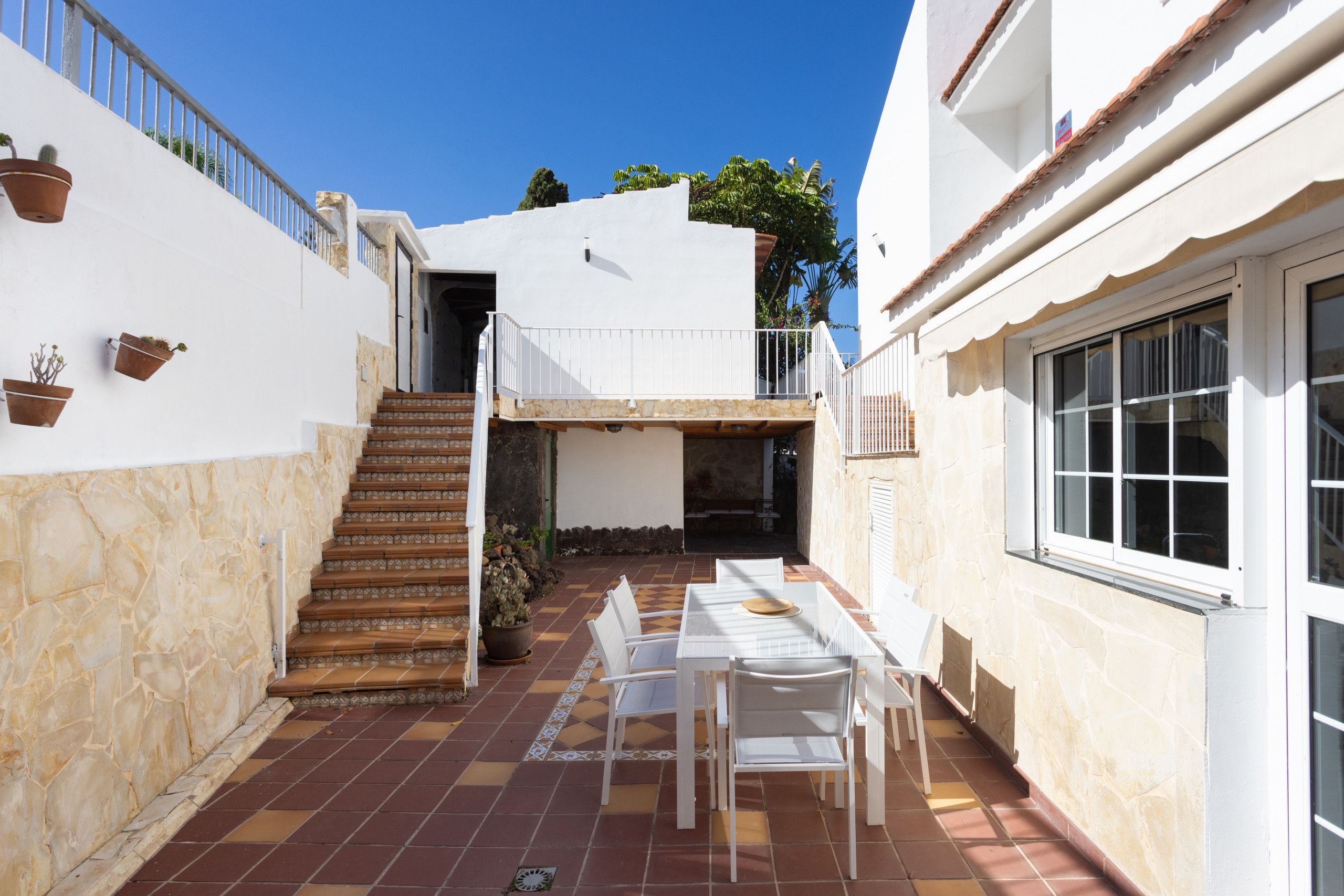 Une cour avec un sol carrelé, un escalier, un petit espace extérieur aménagé avec une table et des chaises, et un bâtiment blanc doté d’un balcon.