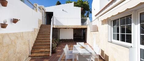 Un patio con suelo de baldosas, una escalera, un pequeño espacio al aire libre con mesa y sillas, y un edificio blanco con balcón.