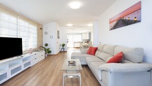 Un salon moderne avec un téléviseur à écran plat, une table basse en verre, un canapé beige agrémenté de coussins rouges et un tableau accroché au mur.