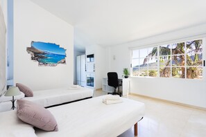 Habitación de hotel con dos camas, un escritorio, una silla y un ventanal con vistas a palmeras y una playa.