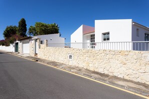 Una calle con un edificio blanco, un muro de piedra y un cielo azul despejado.