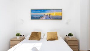 Une chambre à coucher avec un grand lit, deux tables de chevet, un tableau représentant un coucher de soleil sur une jetée, et une petite plante sur chaque table de chevet.