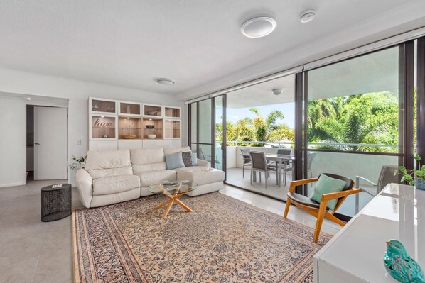 Relax in this spacious living room with a plush sofa, glass coffee table, and dining area, connected to a balcony with a view of lush greenery.