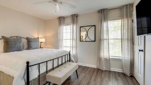 Un dormitorio con cama, un banco, ventilador de techo, ventana con cortinas y espejo montado en la pared.