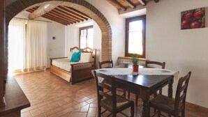 A room with a wooden dining table, chairs, a bed with a blue pillow, a potted plant, and a fruit painting on the wall. - Resort Le Tegole (Riparbella)
