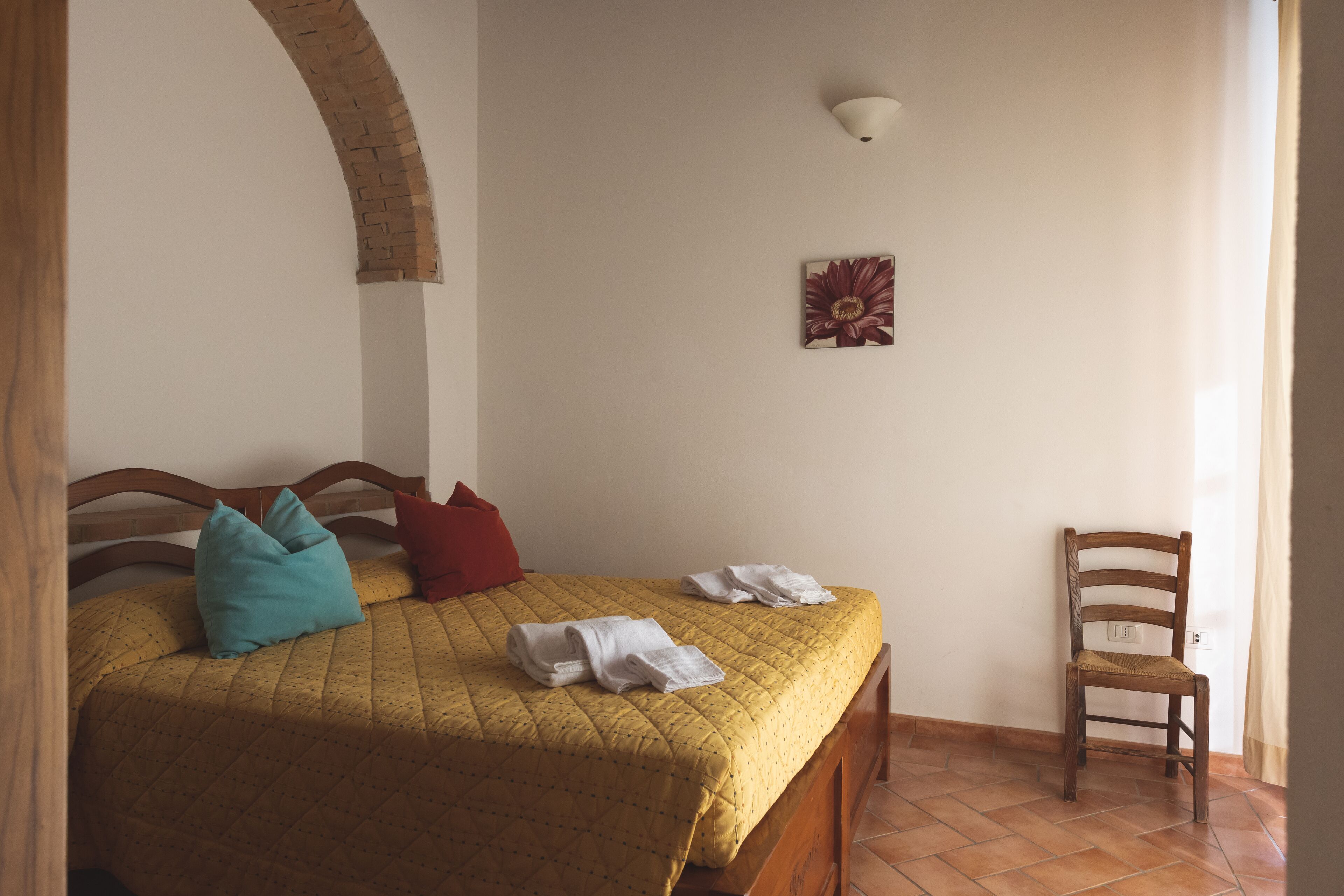 A bed with a quilted cover, two pillows, and folded towels on a wooden bed frame. A wooden chair is positioned against the wall.