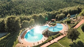 Vista aérea de un resort con dos piscinas circulares rodeadas de sillas de descanso y sombrillas, con un denso bosque de fondo.