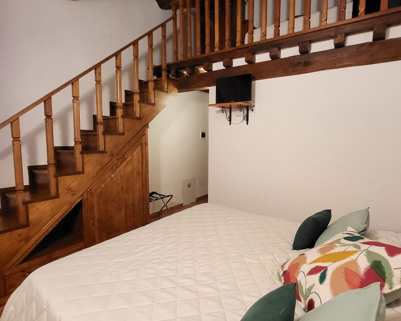 Una camera da letto con una scala in legno, un letto con copripiumino bianco trapuntato e un cuscino colorato con motivi floreali.