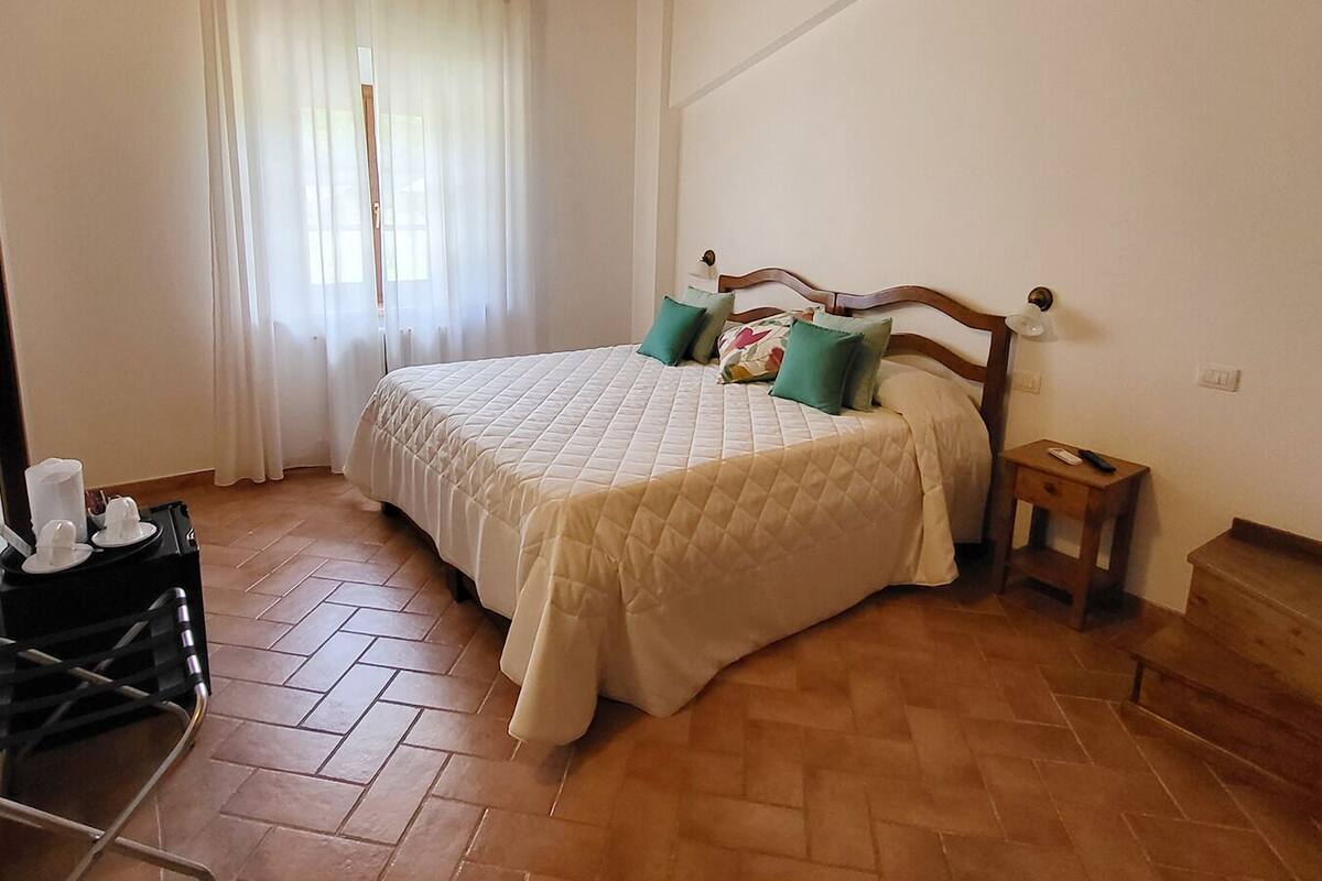A bedroom with a bed, a nightstand, a wooden headboard, a window with curtains, and a tiled floor.