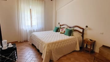 Quarto com cama, mesa de cabeceira, cabeceira de madeira, janela com cortinas e piso ladrilhado.