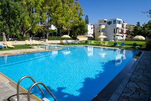 A clear blue swimming pool with lounge chairs and umbrellas, surrounded by trees and a modern building. - Prince of Lillies (Hersonissos)