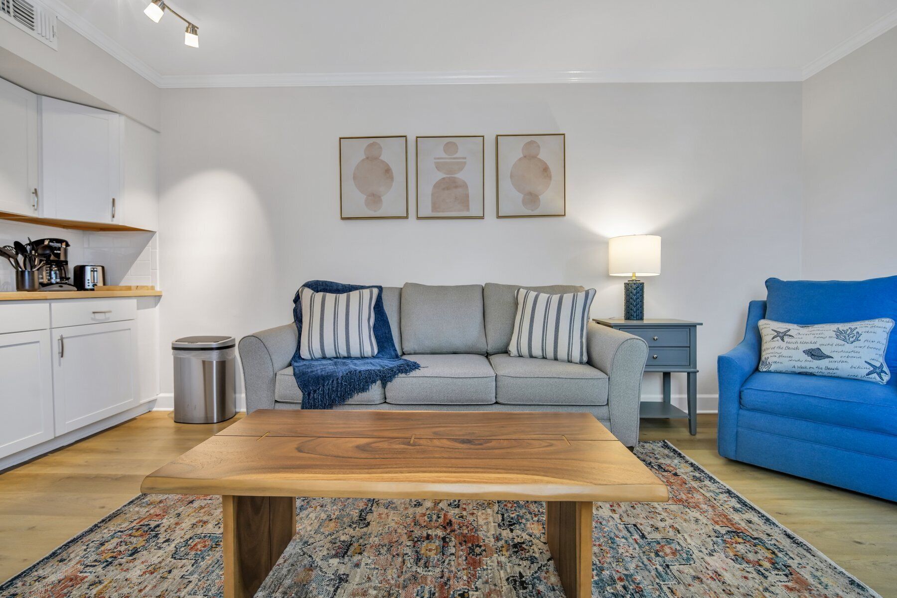 A living room with a wooden coffee table, a grey sofa, a blue armchair, and a patterned rug. There is a small side table with a lamp, a trash bin, and a kitchen area with white cabinets and a coffee machine.