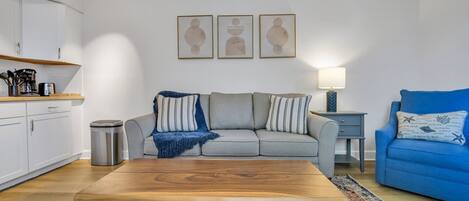A living room with a wooden coffee table, a grey sofa, a blue armchair, and a patterned rug. There is a small side table with a lamp, a trash bin, and a kitchen area with white cabinets and a coffee machine.