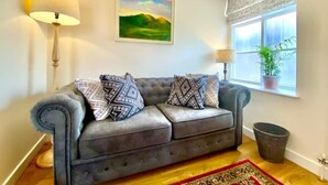A living room with a grey sofa, patterned cushions, a painting, a lamp, a window with a view, and a potted plant. - Tilly's Perfect Apartment in charming Ledbury (Ledbury)