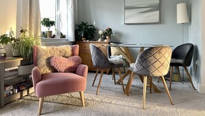 A modern living room with a pink armchair, a glass dining table, and two chairs with wooden legs. There are potted plants on a shelf and a large painting on the wall. - Beautiful Cottage close to the beach w/ sunny garden (Crantock)