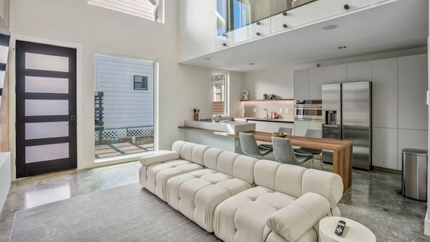 A modern kitchen with stainless steel appliances, a dining area with a wooden table and chairs, and a large beige sofa in a spacious living area with a view of the outside.