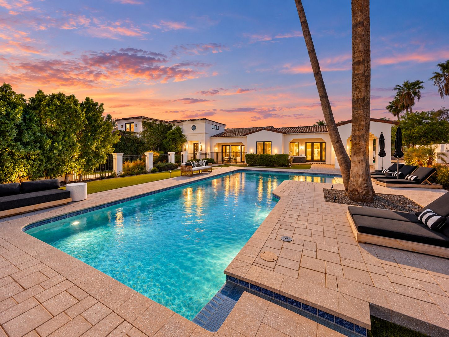 A swimming pool with lounge chairs and a patio area, surrounded by a landscaped garden and a building with multiple windows.
