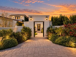 A modern house with a paved driveway leading to a gated entrance, surrounded by well-maintained hedges and flowering plants.