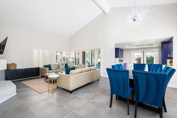 A spacious living area with a sofa, coffee table, and dining table with chairs. There are large windows and a chandelier.