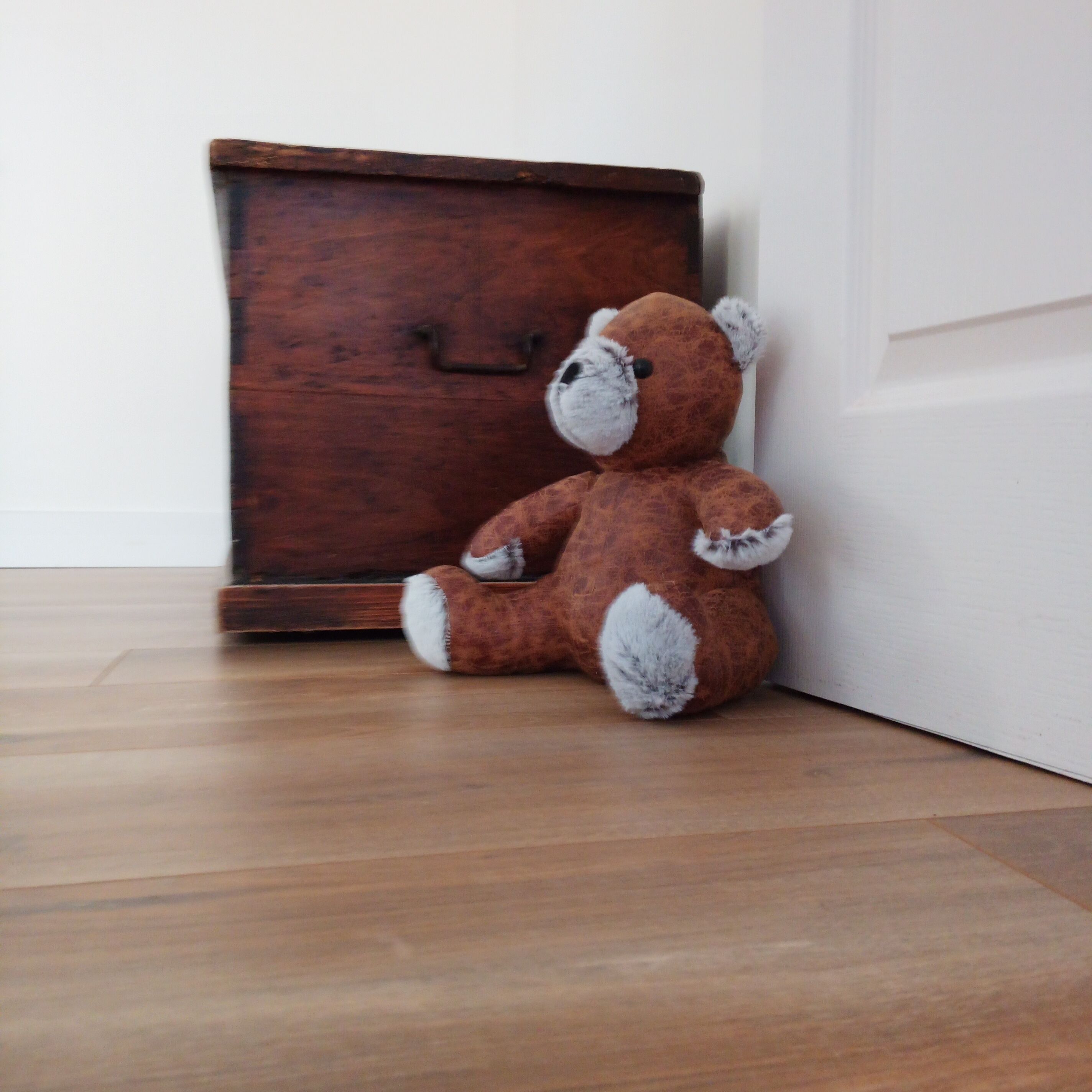 Un ours en peluche marron est assis sur un plancher en bois, à côté d’une armoire en bois.