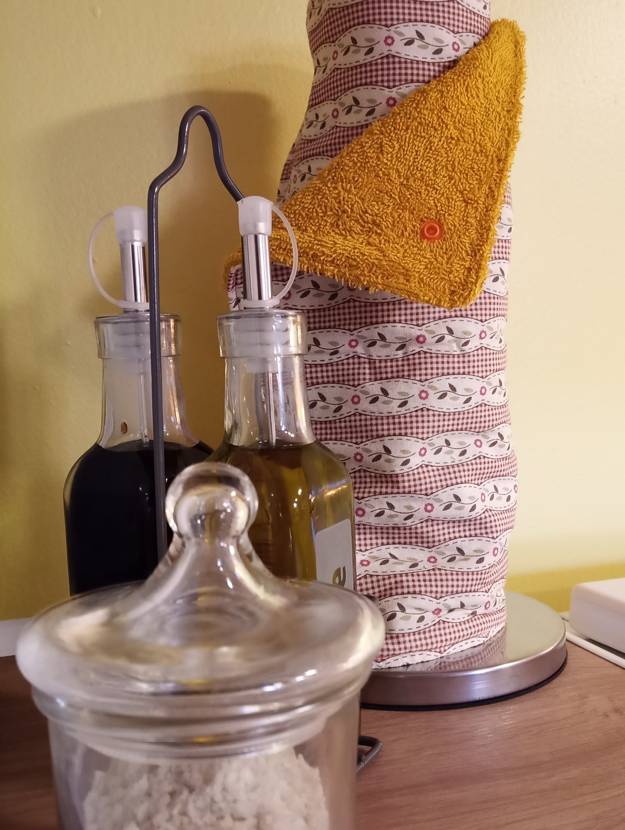 A kitchen counter with a jar of flour, two bottles of olive oil, and a yellow towel hanging on a patterned cloth.