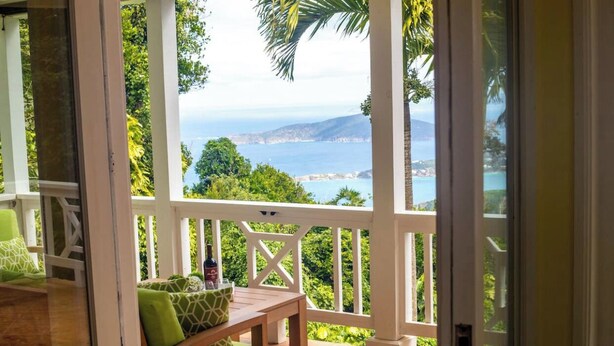Un balcón con mesa de madera y cojines verdes, con vistas a un paisaje costero con árboles y el mar.
