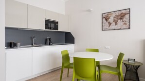 A modern kitchen with white cabinets, a microwave, and a sink. There is a round table with green chairs and a small side table with a clock.