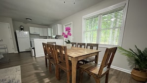 A dining area with a wooden table and chairs, a kitchen in the background, and a window with blinds. - King Suite Hot Tub Fireplace Fire Pit Foosball 1 (Ladson)