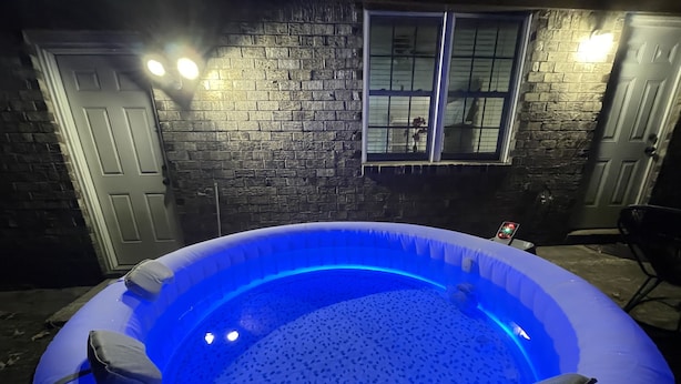An inflatable hot tub with a blue light, surrounded by a brick wall and a window. - King Suite Hot Tub Fireplace Fire Pit Foosball 1 (Ladson)