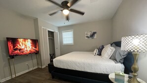 A bedroom with a bed, a TV displaying a fire, a ceiling fan, and a bedside table with a lamp. - King Suite Hot Tub Fireplace Fire Pit Foosball 1 (Ladson)