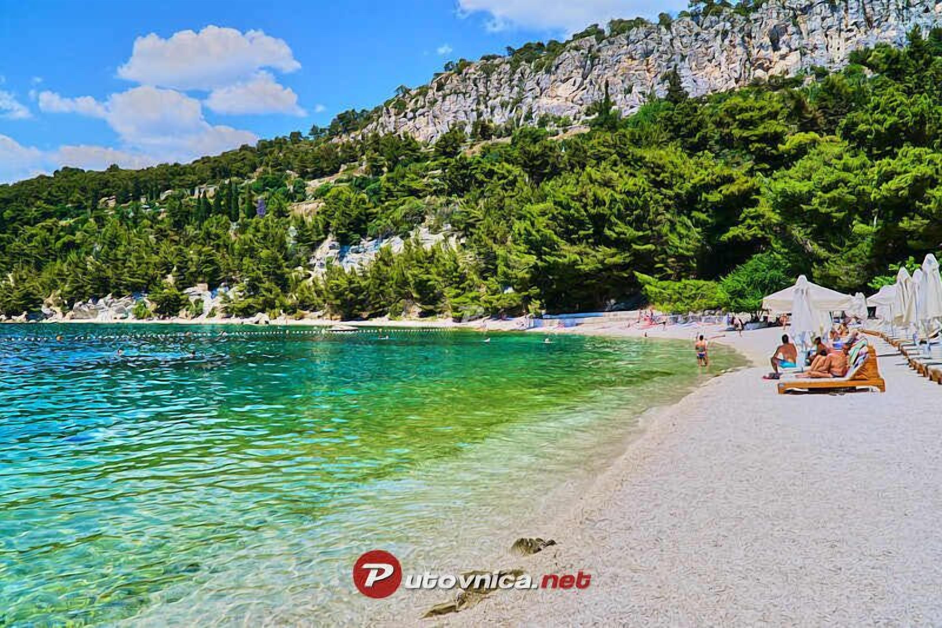 A beach with clear turquoise water, a rocky hillside with trees, and white umbrellas on the sand.